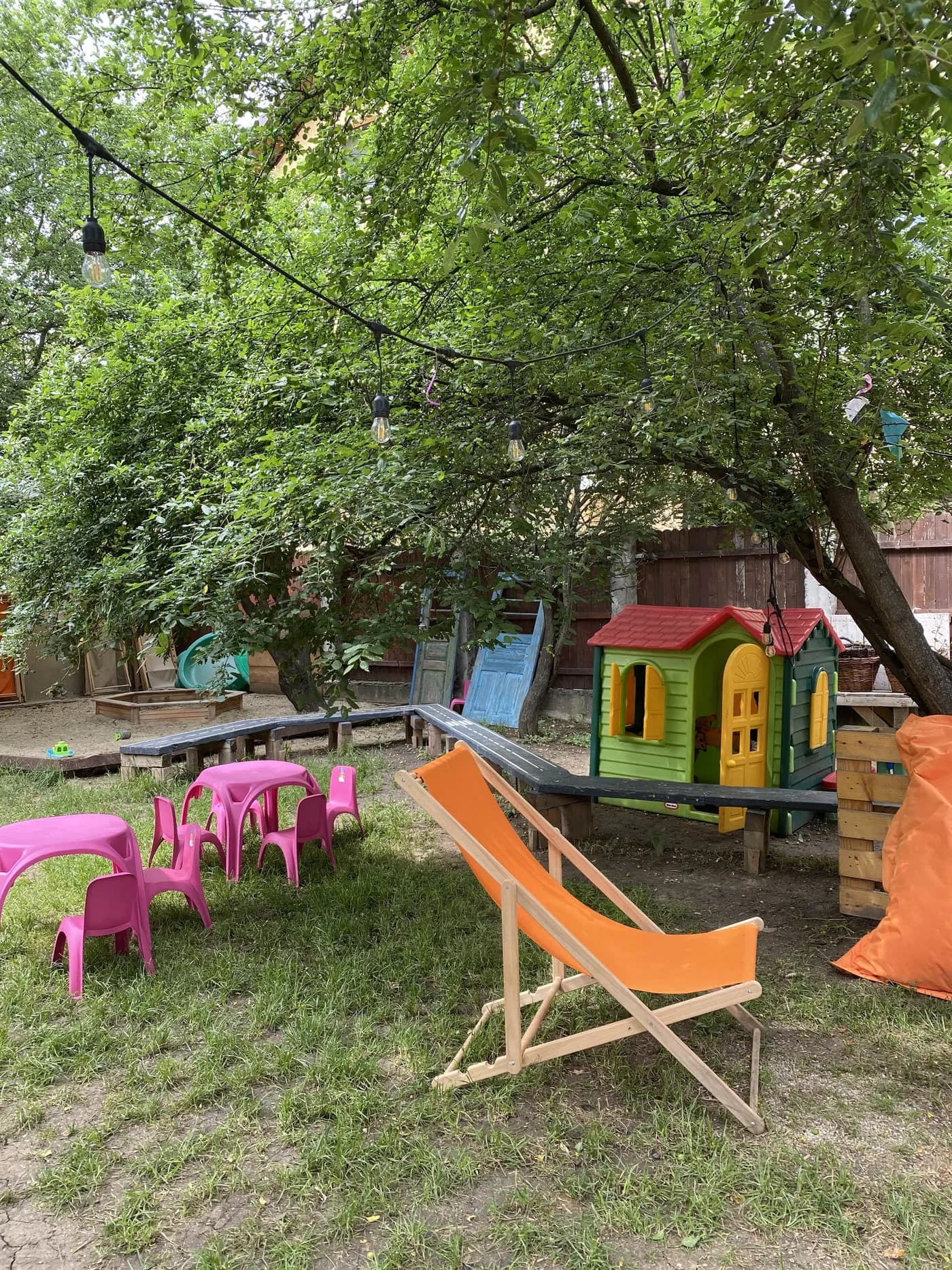 Mese joase, șezlong portocaliu și căsuță de joacă sub coroana unui corcoduș